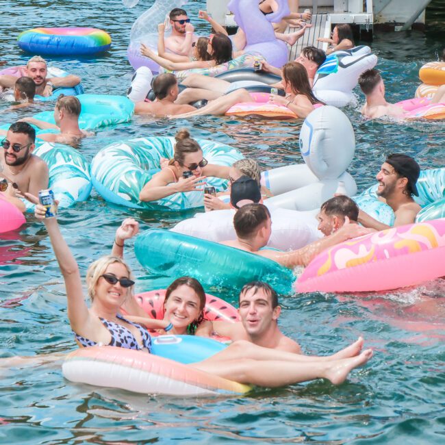 A lively group of people relaxing on colorful inflatable floats in a lake. They are enjoying sunny weather, some holding drinks, and are surrounded by water with a boat in the background. Everyone appears to be having fun.