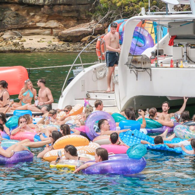 A group of people relaxing on colorful inflatable floats in the water near a white boat. Some are on the boat's deck. The setting is a rocky shoreline with greenery in the background.