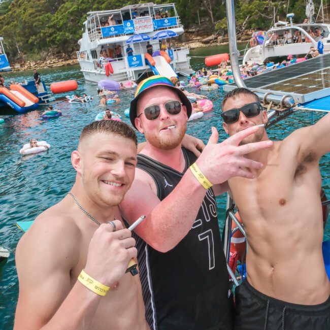 Three men on a boat pose for a photo, smiling and making hand gestures. They are surrounded by water activities and other boats with people enjoying the sunny day. Inflatable toys and swimmers can be seen in the background.