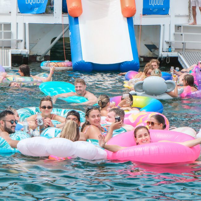 A group of people enjoying a party on inflatable floats in the water near a boat with an inflatable slide. They are smiling, relaxing, and holding drinks on various colorful pool floats.