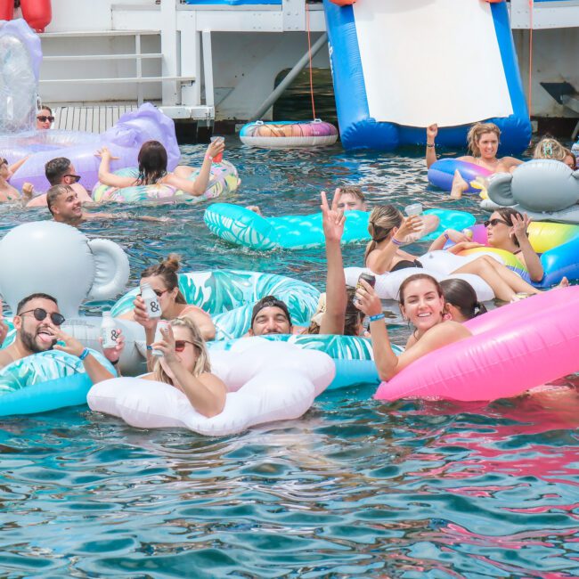 A group of people enjoying a sunny day on a lake, floating on colorful inflatable rafts, including elephants and unicorns. They are smiling and holding drinks, with a large slide in the background attached to a boat.