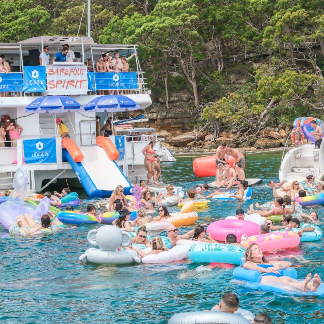 A lively scene of people enjoying a day on the water. A large boat is anchored nearby, and many individuals are floating on colorful inflatable tubes and rafts. Trees line the shore in the background, creating a vibrant summer atmosphere.