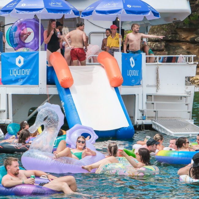 A group of people relax in a scenic waterside setting. Some are on a boat equipped with a slide, while others float nearby on inflatable rings. The water is calm, and the mood is festive and leisurely.