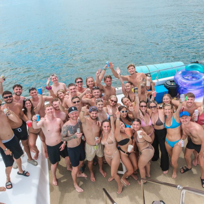 A large group of people in swimwear gather on a boat, smiling and holding drinks. The ocean provides a scenic backdrop. Inflatable tubes are visible on the deck. The sunny atmosphere suggests a joyful summer outing.