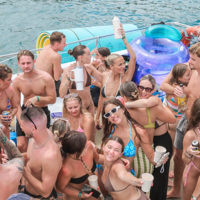 A group of people enjoying a sunny day on a boat, holding drinks and wearing swimwear. Some people are floating on inflatable rings, and everyone appears to be smiling and having fun near the water.