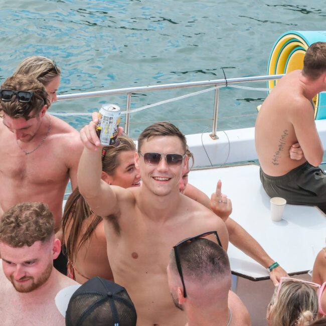 A young man on a boat holds up a can and smiles at the camera. He's surrounded by a group of people enjoying a sunny day, all wearing swimsuits. Behind them, the water is visible, along with pool noodles and a boat railing.