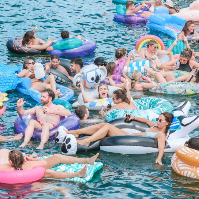 A large group of people relaxing on colorful inflatable rafts in a body of water on a sunny day. Some wear sunglasses, while others chat and enjoy the festive atmosphere. Various raft shapes include a panda, unicorn, and rainbow.