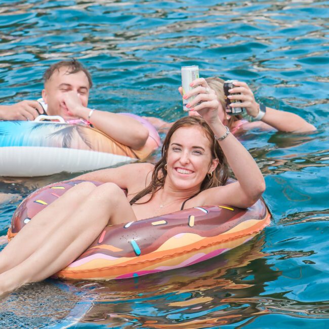 Two people relax in colorful inflatable rings in the water. The woman in front smiles while holding up a drink, and the man behind her appears relaxed with a beverage. The water is clear and blue.