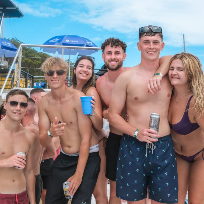 A group of six young adults enjoying a sunny day on a boat, some holding drinks. They're smiling and wearing swimsuits, with a vibrant blue sky and a hint of trees in the background. Other people and boats are visible in the scene.