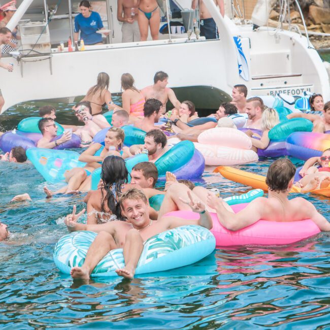 A lively scene of a group of people enjoying a sunny day on colorful inflatable floats in the water near a boat. The boat is anchored, and more people are having fun onboard. The atmosphere is vibrant and relaxed.