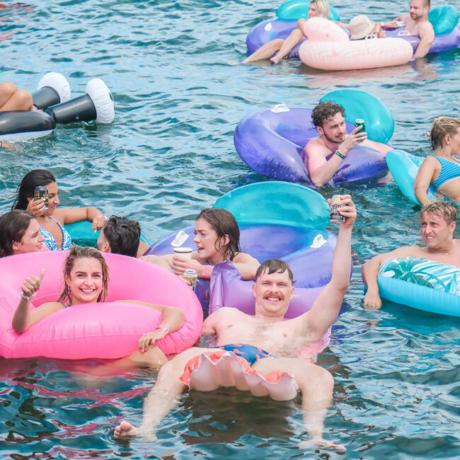 People relaxing in a body of water on colorful inflatable tubes, holding drinks and smiling. The scene is lively, with multiple groups enjoying their time on various floaties, including watermelon and donut designs.