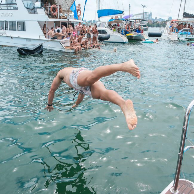 Person diving off a boat into the water, surrounded by several anchored boats and people enjoying a sunny day.