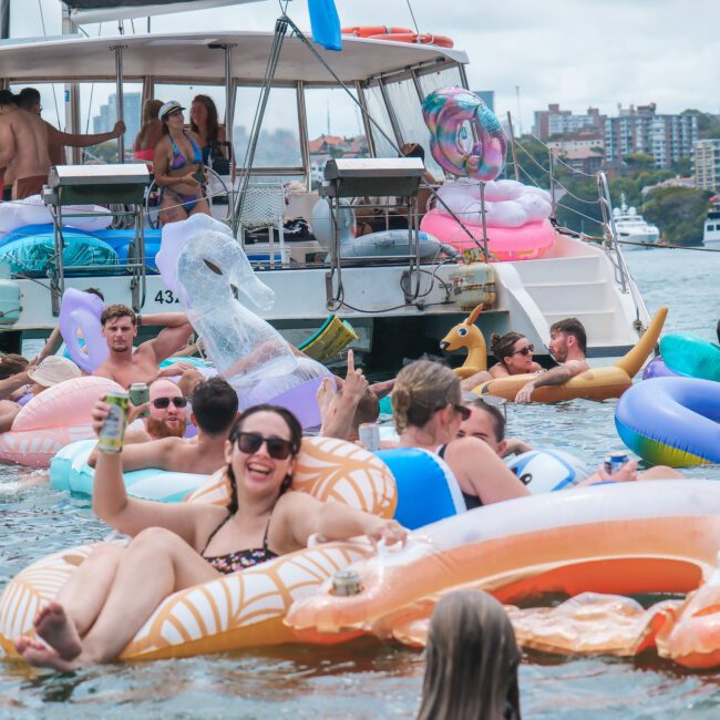 A group of people enjoying a sunny day on the water, floating on colorful inflatable toys near a boat. The scene includes a T-Rex pool float and other playful shapes. The city skyline is visible in the background.