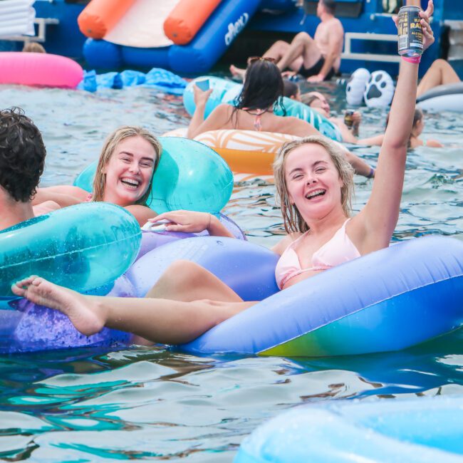 People enjoying a pool party, lounging on colorful inflatable tubes. A smiling woman raises a can in the air. The atmosphere is lively with others swimming and relaxing in the background.