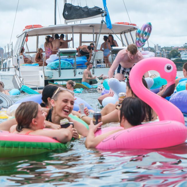 A group of people enjoy a lively pool party by the water, surrounded by colorful inflatable floats, including a pink flamingo. A boat with more partygoers is anchored nearby, with city buildings visible in the cloudy background.