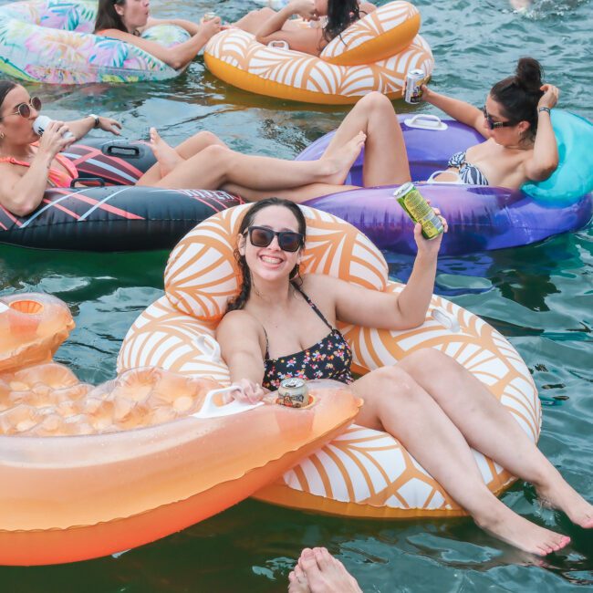 A group of people relax on colorful inflatable tubes in the water. They are wearing swimsuits and sunglasses, enjoying drinks under the sun. The scene conveys a fun and leisurely atmosphere.