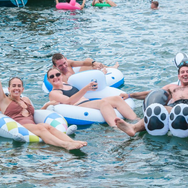 A group of four people relax on inflatable floats in a body of water. They are smiling and two are holding drinks. The floats are colorful and variously shaped, including a bear print design. Other people are visible in the background.