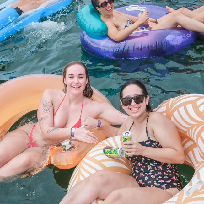 Two women relax on inflatable rafts in a lake, smiling and holding drinks. Others float nearby on various colorful inflatables, enjoying the water and sunshine.