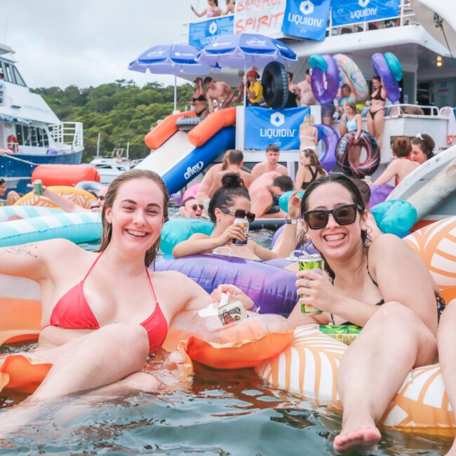 Two people in swimsuits enjoy lounging on inflatable floaties in the water, holding drinks. In the background, a lively crowd is seen on a boat, with more inflatable toys and lively decorations.