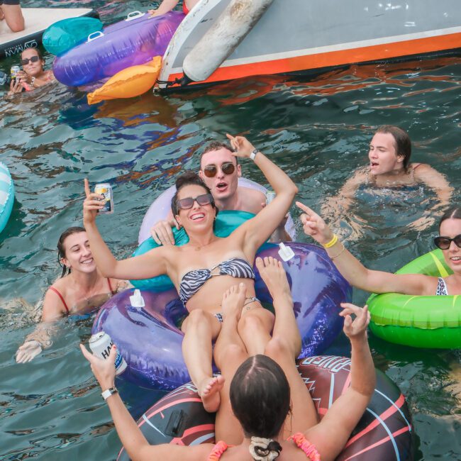 A group of people enjoying a day in the water. One person is lounging on a colorful inflatable, surrounded by friends in swimwear. They are smiling and holding drinks, with boats and other inflatables nearby. The atmosphere is relaxed and cheerful.