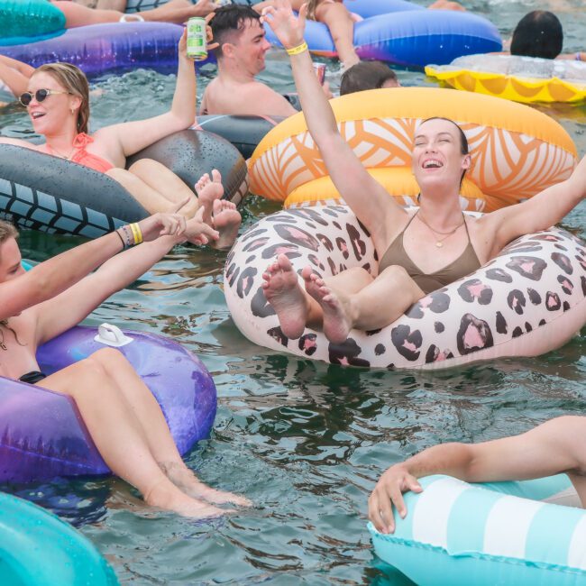 A group of people relaxing and having fun in the water on colorful inflatable tubes. They are smiling and holding drinks, with some raising their arms joyfully. The scene conveys a vibrant and festive summer atmosphere.