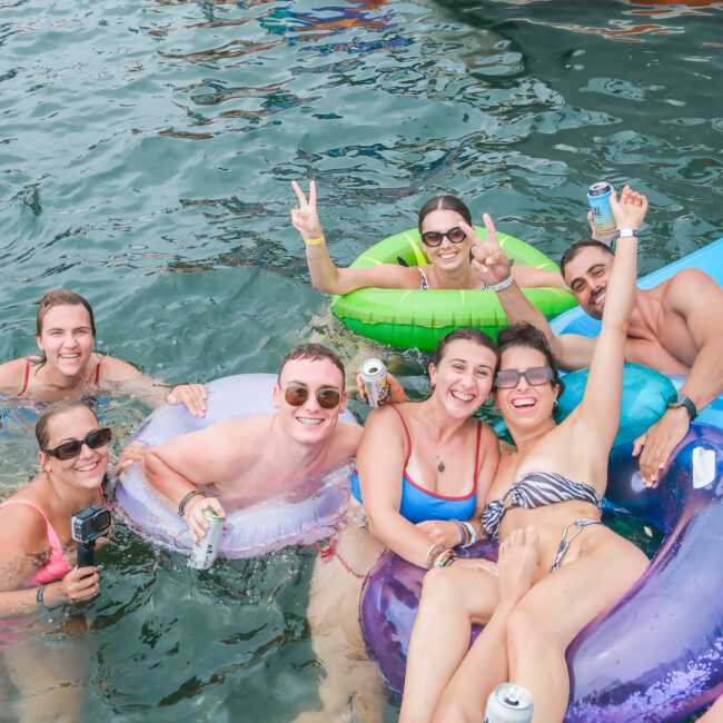 A group of seven people enjoying a day in the water with inflatable floats and drinks. They are smiling and posing for the photo, with one person flashing peace signs.