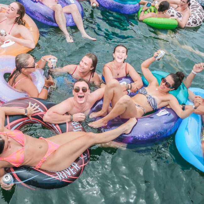 A group of people relaxing and laughing on colorful inflatable floaties in a body of water. They are holding drinks and appear to be enjoying a sunny day. Some floaties are shaped like animals and others are patterns.