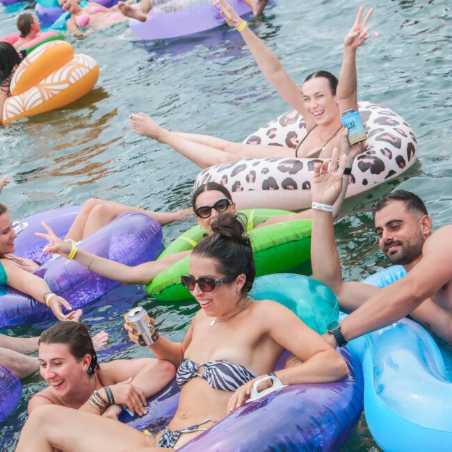 A group of people relaxing and having fun on colorful pool floats in the water. They are smiling, holding drinks, and making peace signs, enjoying a sunny day.