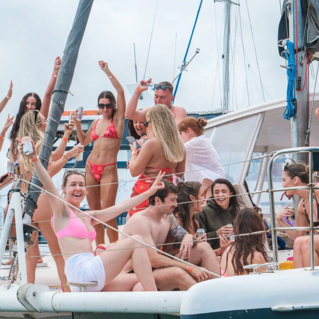 A group of people enjoying a party on a sailboat. They are smiling, dancing, and holding drinks, dressed in casual and swimwear under a cloudy sky. The sailboat is docked, with a partial view of the marina in the background.