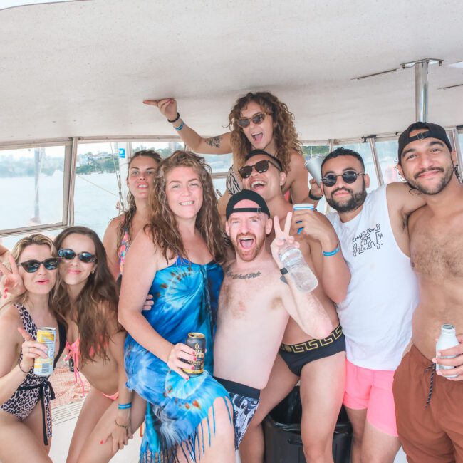 A group of people enjoying a boat party, some in swimsuits and sunglasses. They are smiling, holding drinks, and making peace signs. The atmosphere is relaxed and festive, with the ocean visible in the background.