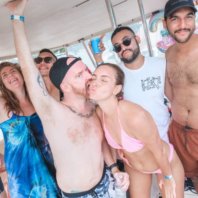 A group of people on a boat, some wearing swimsuits and sunglasses. One person is kissing another on the cheek, while others smile and pose. They're enjoying a sunny day, and a few hold drinks. The background shows water and a cityscape.