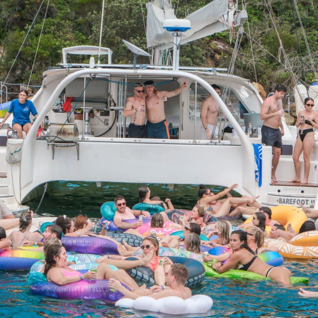 A large group of people enjoying a sunny day on the water with floaties near a catamaran labeled "Barefoot." Some are on the boat, while others relax in the water surrounded by greenery. Everyone seems to be having a good time.