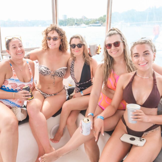 Five women in swimwear sit together on a boat, smiling and holding drinks. The scene is bright, with sunlight coming through windows. The background shows a waterfront with blurred views of trees and buildings.