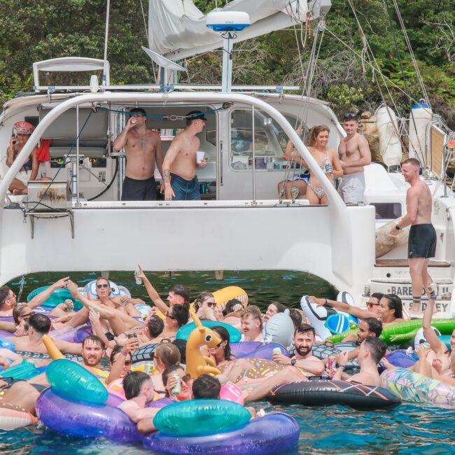 A large group of people are enjoying a sunny day on the water, floating on colorful inflatables around a docked boat. The scene is lively, with some people on the boat and others swimming or resting on the inflatables.