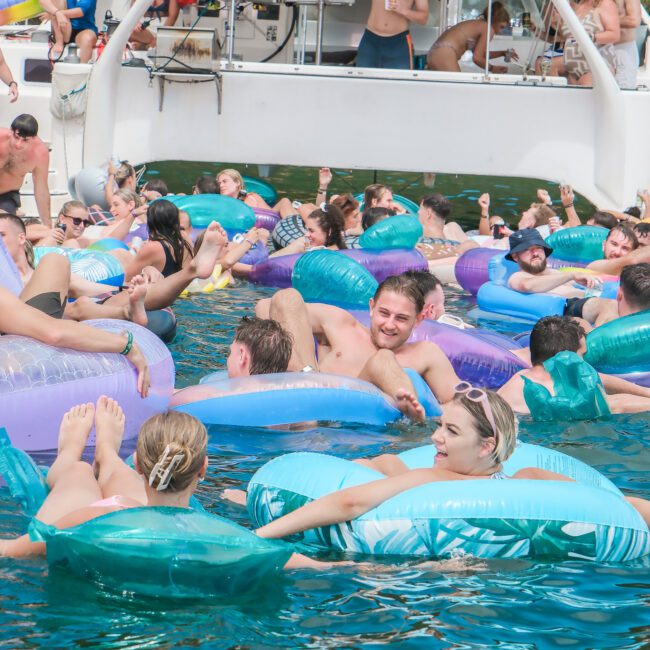 A lively scene of people floating on inflatable tubes in a crowded swimming area near a docked boat. The water is clear, and the inflatables are colorful, with many participants enjoying a sunny day.