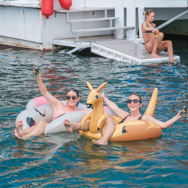 Two people are lounging on animal-themed inflatables in the water, holding drinks with arms raised. A woman in a bikini is seated on a dock ladder in the background near a boat. The scene is lively with a summer vibe.