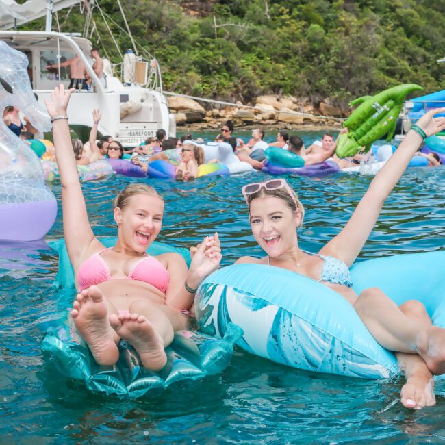 Two women smiling and relaxing on inflatable floats in the water, surrounded by other people enjoying a day out with boats and inflatable toys. Lush greenery and rocky hills are visible in the background under a partly cloudy sky.