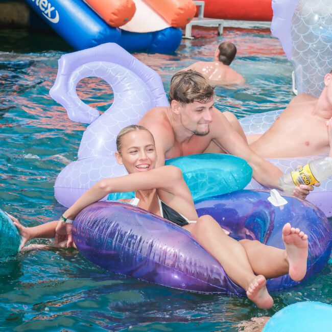 A group of young adults are floating in a pool on inflatable rafts shaped like swans and ducks. They appear to be enjoying a sunny day, laughing and holding drinks. The water is clear, and other pool toys are visible in the background.