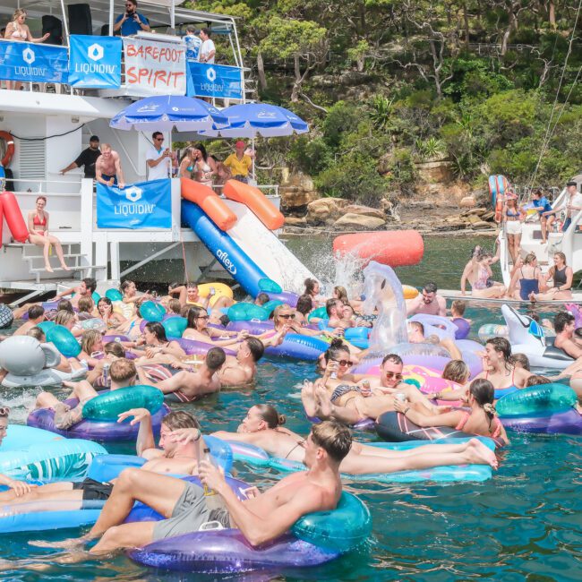 A lively gathering of people enjoying a sunny day on colorful inflatable pool floats in the water, near a boat with a slide. The backdrop includes a wooded shoreline, and participants appear relaxed and joyful.