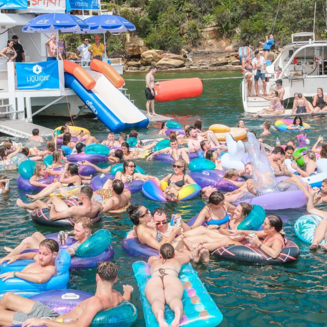 A lively gathering of people enjoying a sunny day on the water. Many are floating on colorful inflatables near boats, with others relaxing or playing. The backdrop features greenery and rocky terrain.