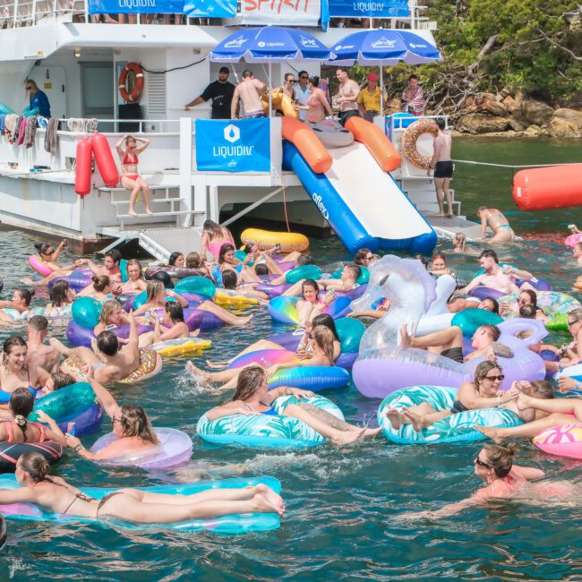 A crowded pool party in the sea with people on colorful inflatables, some shaped like flamingos and unicorns. A white boat anchored nearby has a slide leading into the water. People are enjoying the sunny day.