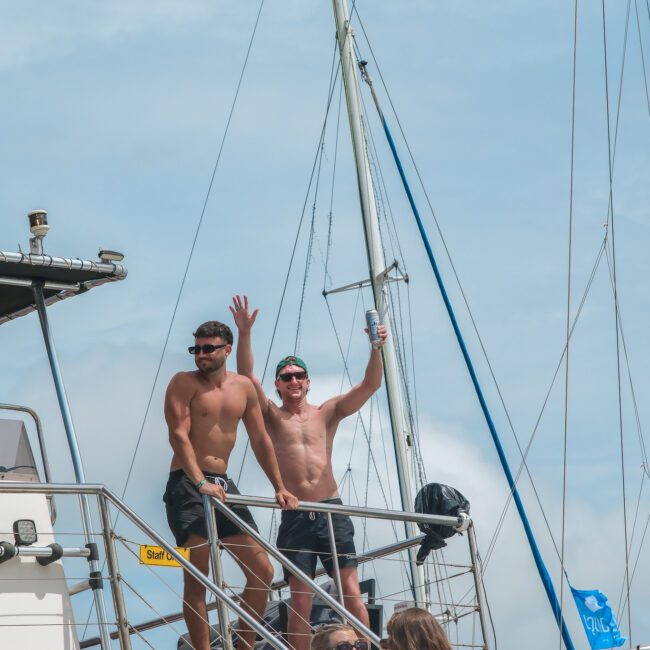 Two shirtless men stand on a boat deck, smiling and waving. One holds a drink while wearing sunglasses. The boat is surrounded by clear skies and has rigging visible. Other people are partially visible in the foreground.