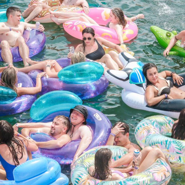 A group of people relaxing on colorful inflatable floats in a body of water. They're enjoying drinks and the sunny weather. The scene is lively, with a mixture of laughter and relaxation.