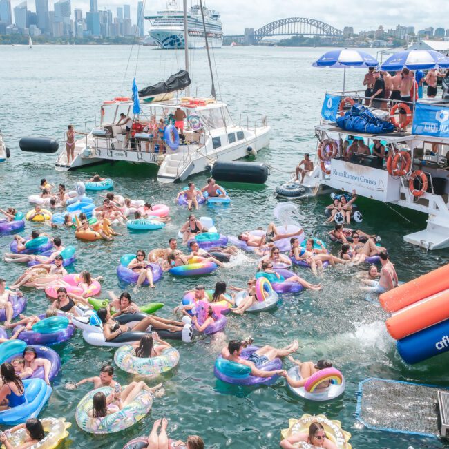 People are enjoying a party on a sunny day in a harbor filled with colorful inflatable rings. Boats and a waterslide into the water add to the festive atmosphere. A city skyline and bridge are visible in the background.