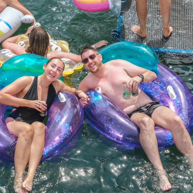 Two people are lounging on bright inflatable chairs in the water, smiling and holding drinks. They are part of a group enjoying a day near a dock, with more fun inflatables and colorful floats in the background.