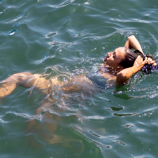 A person floating and relaxing on their back in the water, with arms above their head, eyes closed, and a calm expression. The water appears greenish and sunlit. A logo reading "Yacht Beach" is in the lower right corner.