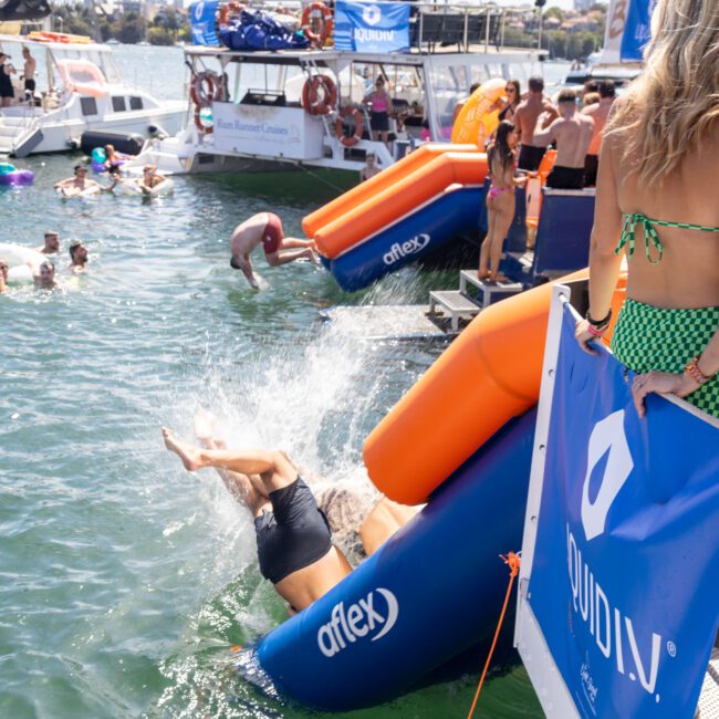 People enjoying a sunny day at a waterfront event. A person is sliding into the water from a colorful waterslide attached to a boat. Others are swimming or floating with inflatables nearby. Boats and more people are visible in the background.