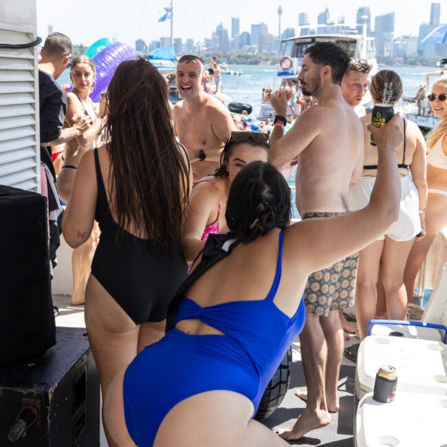 A group of people in swimwear dance and enjoy themselves on a boat. The city skyline is visible in the background. The atmosphere is lively and festive, with laughter and socializing.