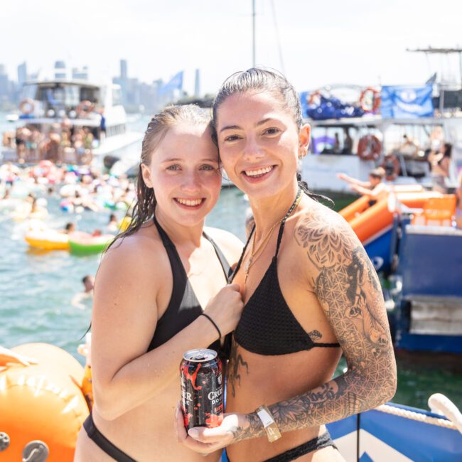 Two women in swimsuits smiling at the camera on a boat party. One holds a can of Coca-Cola. The background shows a crowded scene with people on inflatables and boats on the water. The city skyline is visible in the distance under a sunny sky.