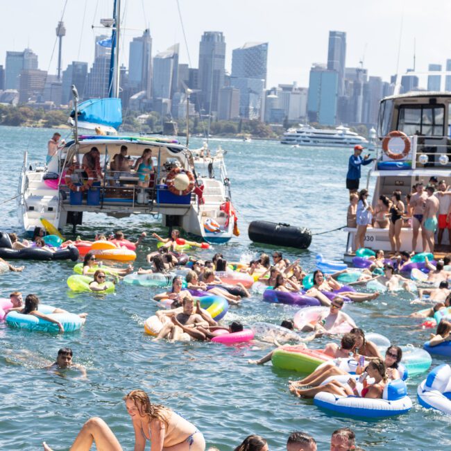 A group of people enjoying a sunny day on colorful inflatables in a harbor, with boats and the city skyline in the background.